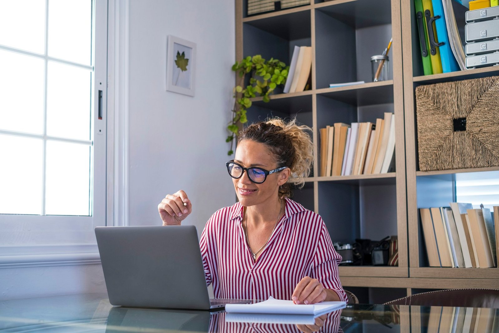 Smiling adult female looking at laptop screen reading email or content and writing notes at home